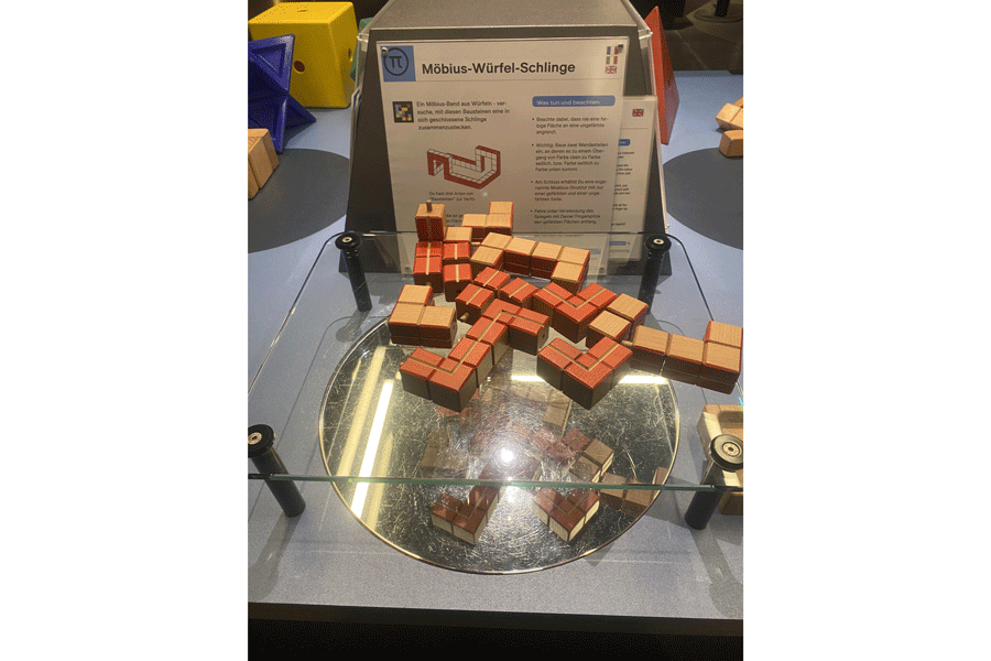 Auf einer Glasplatte, die durch vier Stützen in den Ecken ungefähr 15 Zentimeter über dem Tisch festgemacht ist, liegen viele Holzwürfel. Diese haben holzfarbige und rote Seiten. In den roten Seiten gibt es eine Einkerbung. Die Würfel sind in kleinen Einheiten zusammengesteckt. Hinten an den Würfeln hängt die Anleitung. Auf der An-leitung ist ein NaviLens-Code angebracht. Er sieht aus wie einfarbiger QR-Code.
