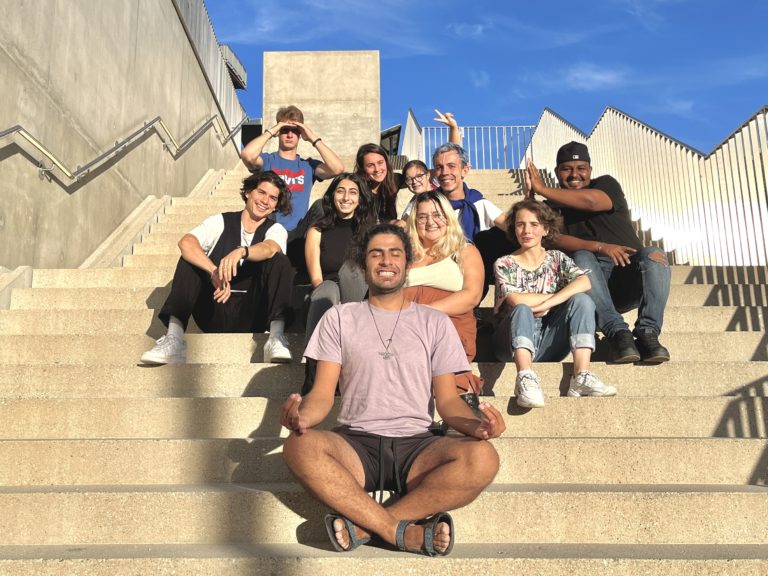 Un escalier extérieur beige sur fond de ciel bleu. Dix jeunes personnes souriantes, aux tenues estivales, sont assises sur l’escalier. Le jeune homme au premier plan croise les pieds et pose ses mains sur ses cuisses, comme s’il méditait.