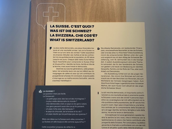 Un panneau avec trois blocs de textes noirs sur fond blanc et un texte blanc sur fond noir. Le titre « La Suisse. C’est quoi ? » est écrit en français, allemand, italien et anglais. En haut du panneau, le contour d’un écusson avec une croix.