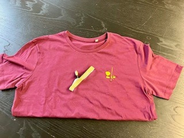 Ein T-Shirt in bordeauxrot mit einer gelben Stickerei. Die Stickerei zeigt abstrakt drei Steine und einen Holz-stab. Auf dem T-Shit liegt ein goldfarbiges Armband mit dem Schriftzug Förderbar.